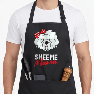 Sheepdog english aprons - Cartoon white dog head with red polka dot bow and SHEEPIE Mama text — shown on a black cotton canvas apron against a white background
