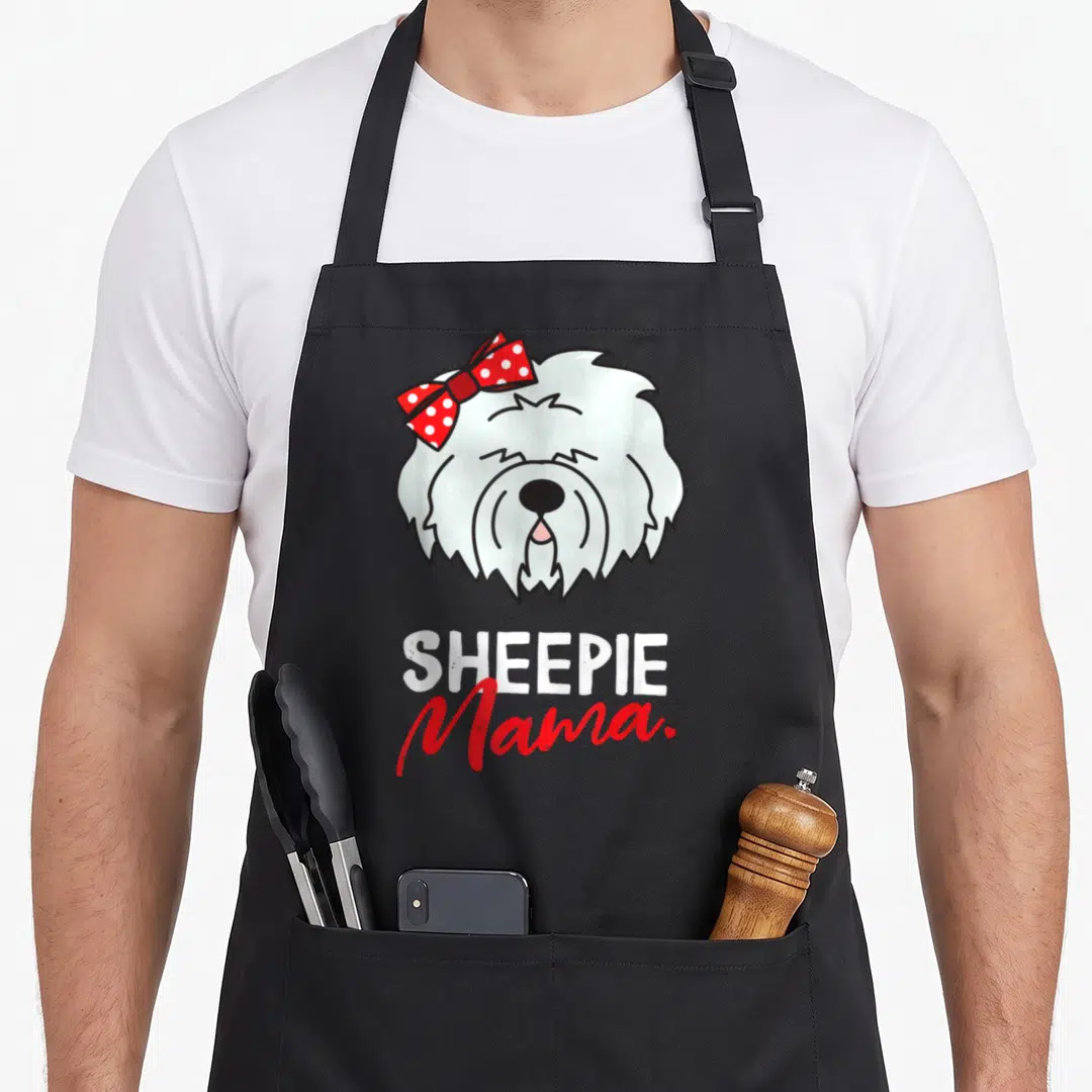 Sheepdog english aprons - Cartoon white dog head with red polka dot bow and SHEEPIE Mama text — shown on a black cotton canvas apron against a white background