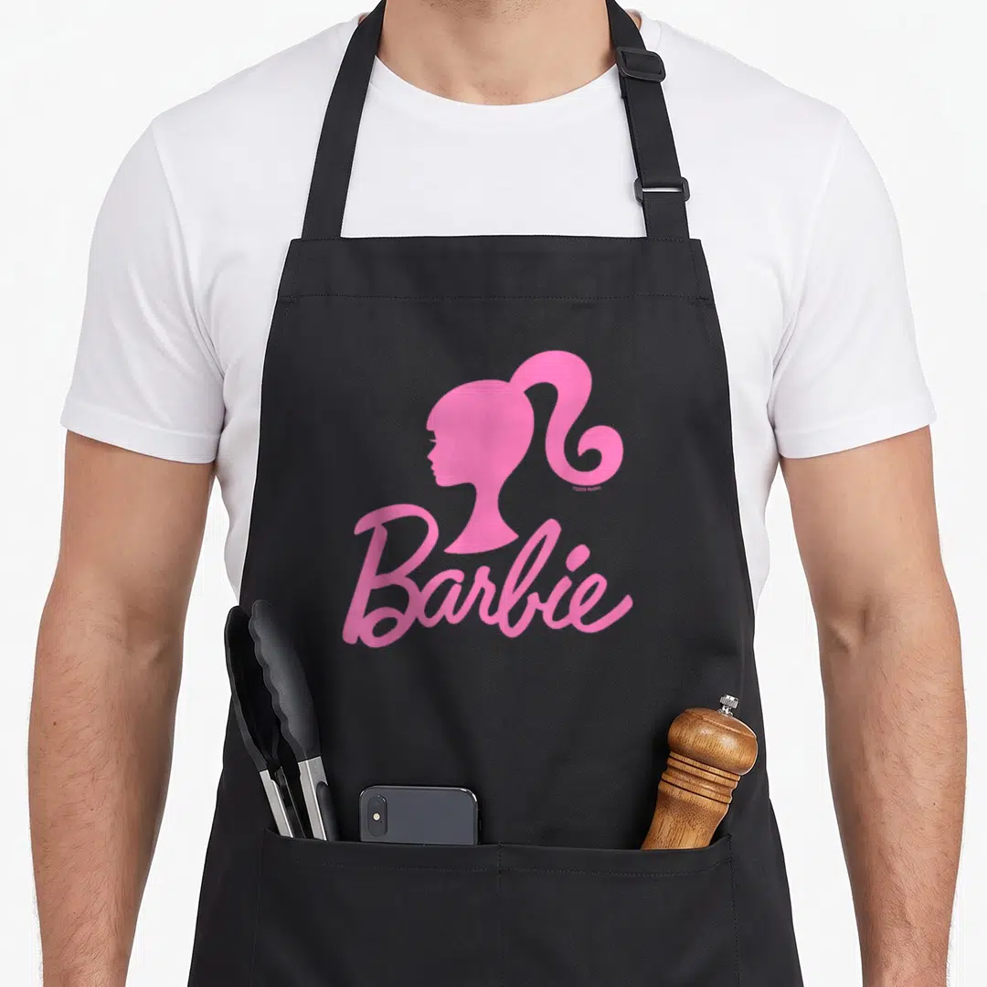 Cool general apron - Pink Barbie head silhouette above cursive Barbie text, minimalist design — shown on a black cotton canvas apron against a white background