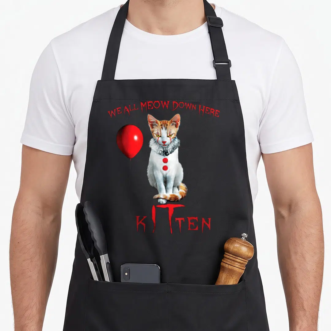 Meow clown aprons - Realistic clown cat with red balloon and WE ALL MEOW DOWN HERE text — shown on a black cotton canvas apron against a white background