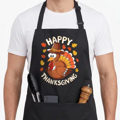 Family dinner thanksgiving apron - Cartoon brown turkey in a hat with HAPPY THANKSGIVING text and autumn leaves — shown on a black cotton canvas apron against a white background