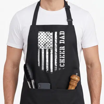 Cheerleader father day apron - Distressed white USA flag and vertical CHEER DAD text on black apron — shown on a black cotton canvas apron against a white background