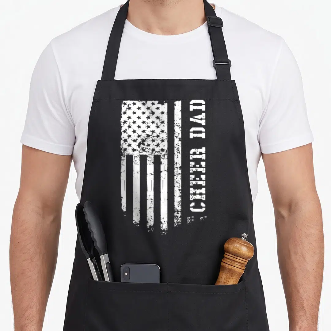 Cheerleader father day apron - Distressed white USA flag and vertical CHEER DAD text on black apron — shown on a black cotton canvas apron against a white background