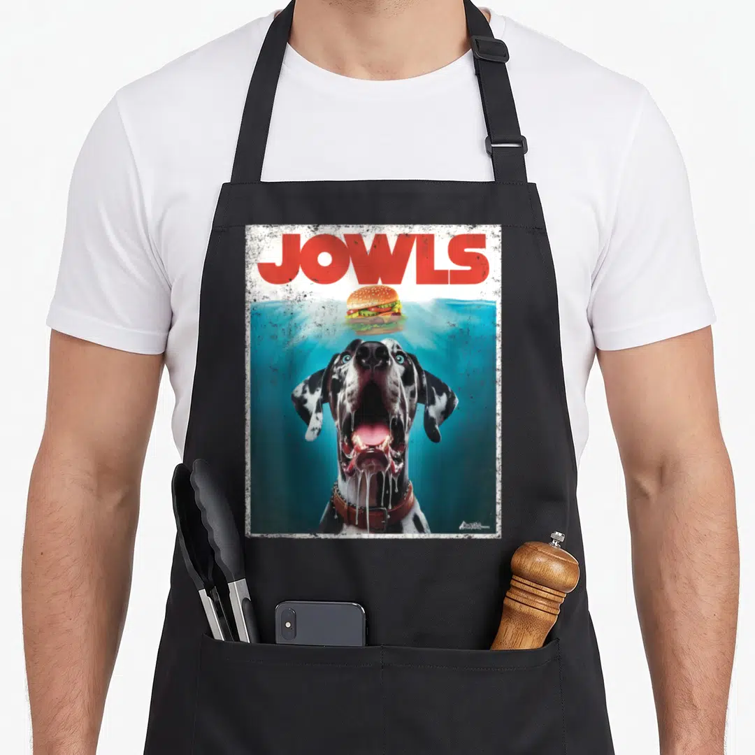 Harlequin dog apron - Realistic black and white dog with blue eyes looking at a floating hamburger under red JOWLS text — shown on a black cotton canvas apron against a white background
