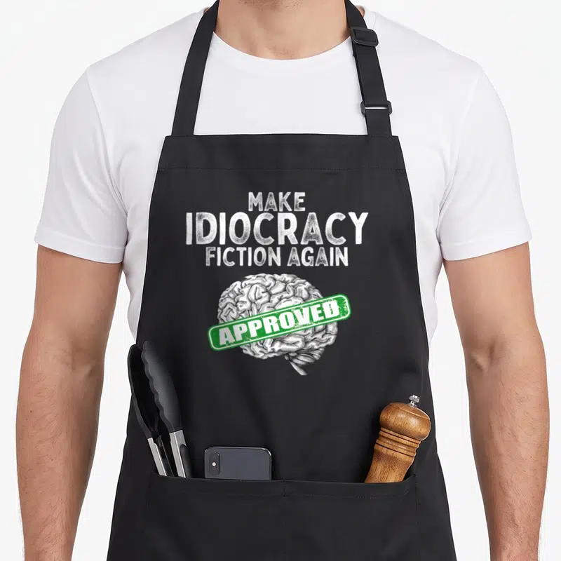 Idiocracy fiction apron - Distressed white text 'Make Idiocracy Fiction Again' above a brain with a green approved stamp — shown on a black cotton canvas apron against a white background