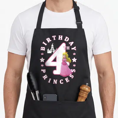 4th princess apron - Princess Peach standing before a castle inside a pink and white circular birthday text design — shown on a black cotton canvas apron against a white background