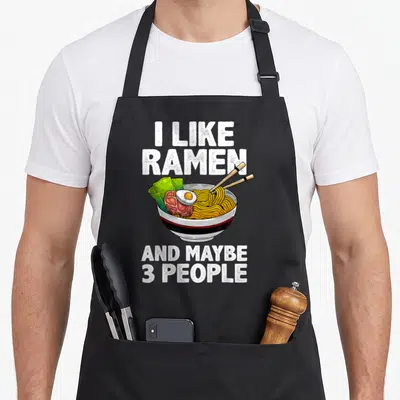 Soup anime apron - Cartoon ramen bowl with chopsticks and text I like ramen and maybe 3 people in white font — shown on a black cotton canvas apron against a white background