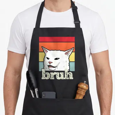 Greeting cat apron - Cartoon white cat over red orange yellow blue-grey stripes with bruh text — shown on a black cotton canvas apron against a white background