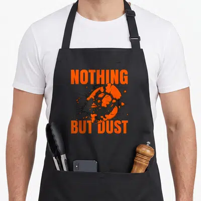 Dust nothing but apron - Orange sphere exploding into black shards between orange NOTHING BUT DUST text — shown on a black cotton canvas apron against a white background