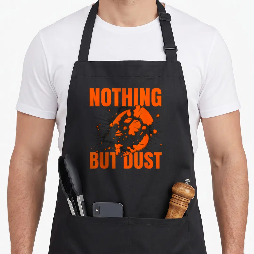 Dust nothing but apron - Orange sphere exploding into black shards between orange NOTHING BUT DUST text — shown on a black cotton canvas apron against a white background