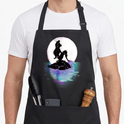 Ariel colorful aprons - Minimalist mermaid silhouette on rock with large moon and blue water reflections — shown on a black cotton canvas apron against a white background