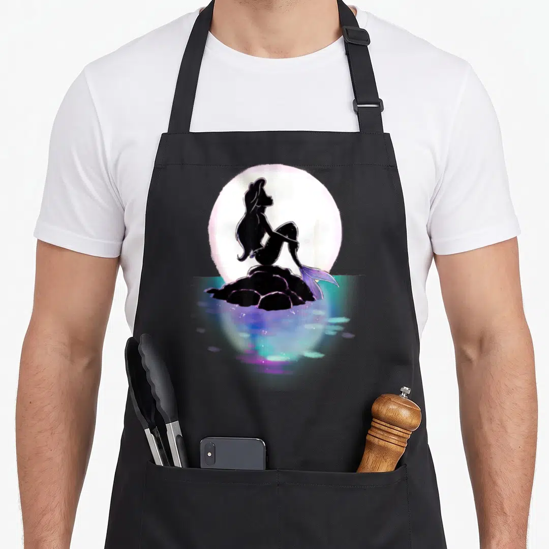 Ariel colorful aprons - Minimalist mermaid silhouette on rock with large moon and blue water reflections — shown on a black cotton canvas apron against a white background