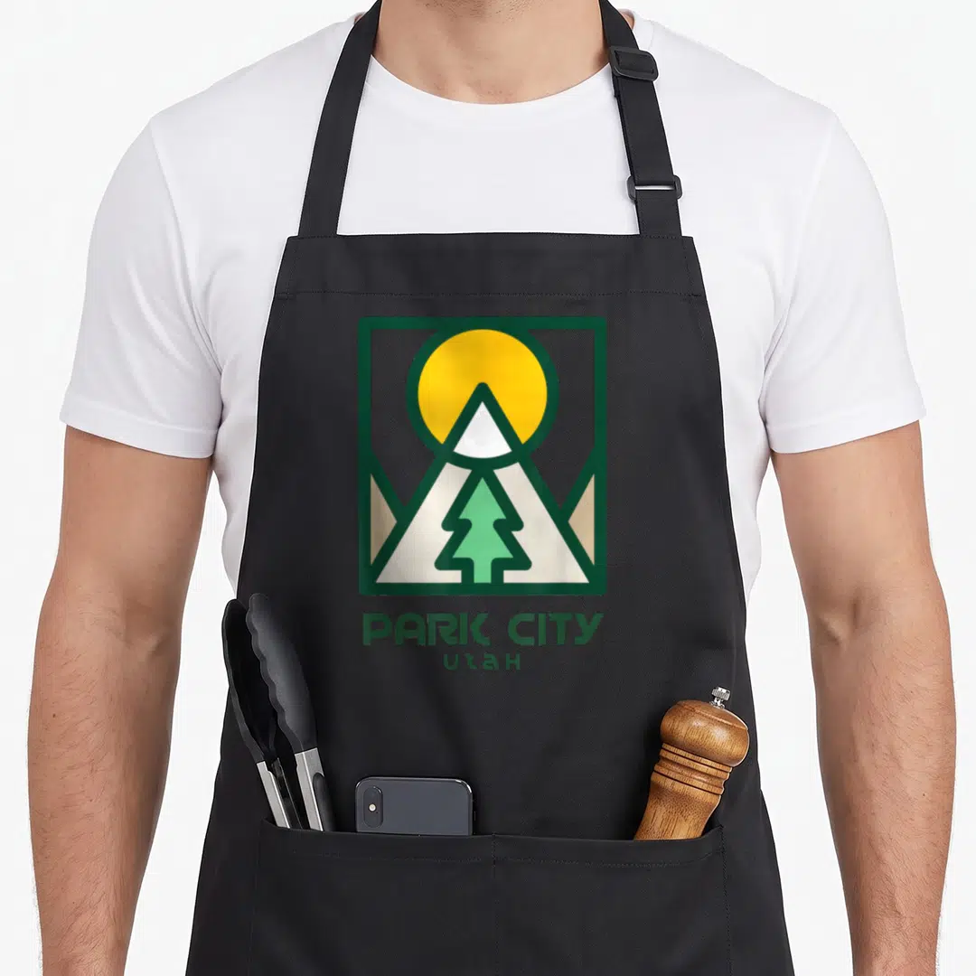 Utah tree apron - Minimalist square logo with yellow sun, white mountain, green pine tree and text PARK CITY UTAH — shown on a black cotton canvas apron against a white background