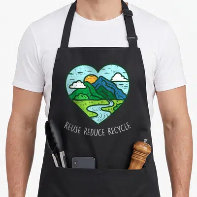 Reuse earth apron - Heart shaped landscape with REUSE REDUCE RECYCLE text in white arch — shown on a black cotton canvas apron against a white background