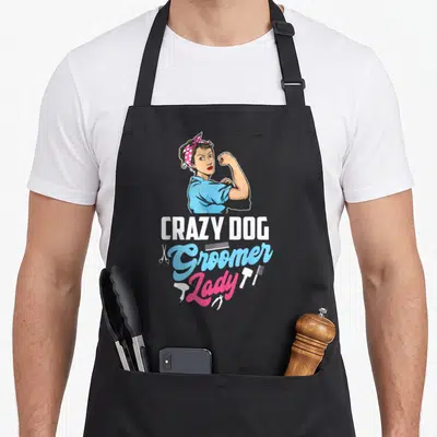 Grazy dog groomer apron - Cartoon woman flexing arm above Crazy Dog Groomer Lady text with scissors and combs — shown on a black cotton canvas apron against a white background