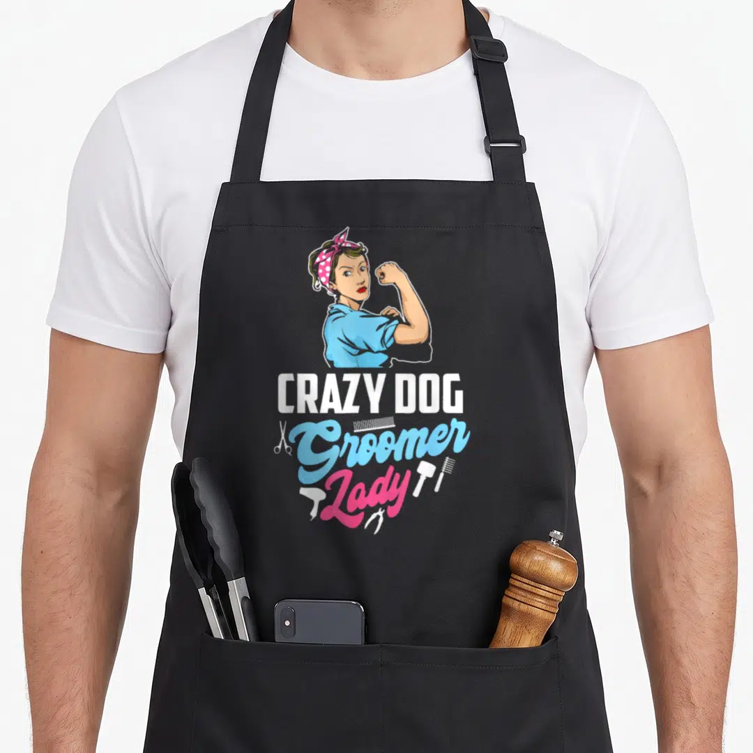 Grazy dog groomer apron - Cartoon woman flexing arm above Crazy Dog Groomer Lady text with scissors and combs — shown on a black cotton canvas apron against a white background
