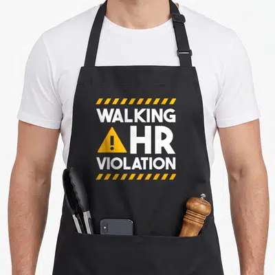 Human walking apron - WALKING HR VIOLATION text with yellow warning triangle and hazard stripes on grey — shown on a black cotton canvas apron against a white background