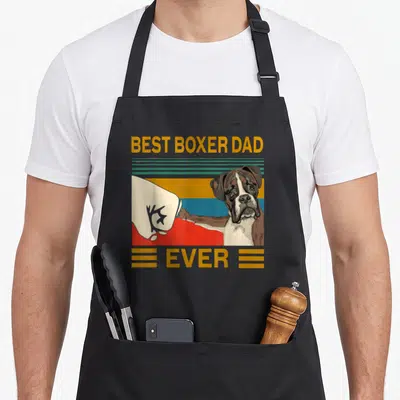 Bump boxer dog apron - Cartoon boxer dog fist bumping glove with BEST BOXER DAD EVER text on striped background — shown on a black cotton canvas apron against a white background