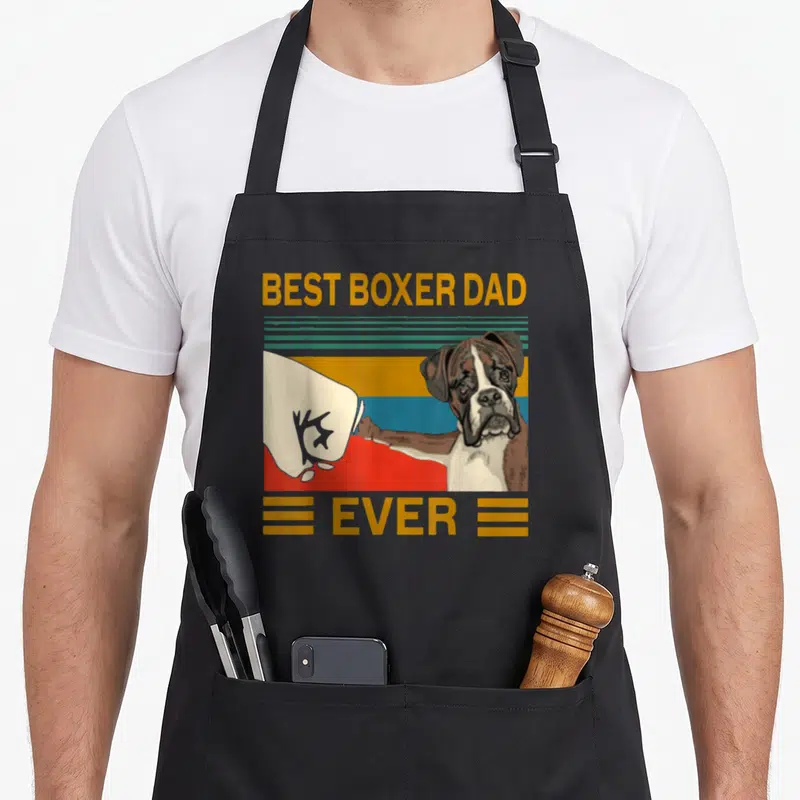 Bump boxer dog apron - Cartoon boxer dog fist bumping glove with BEST BOXER DAD EVER text on striped background — shown on a black cotton canvas apron against a white background