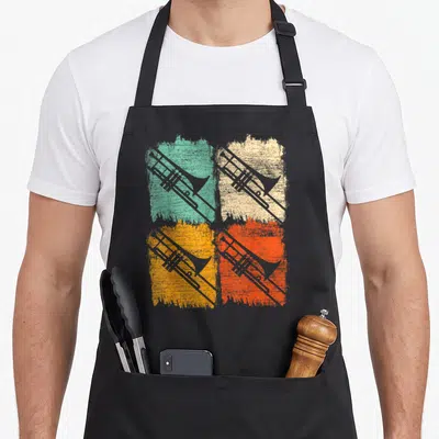Funny trombone quotes apron - Four grey trombones on distressed teal, beige, yellow, and orange paint splashes — shown on a black cotton canvas apron against a white background
