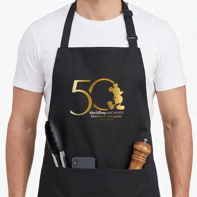 Walt magic apron - Gold 50 with Mickey silhouette and 1970-2020 text — shown on a black cotton canvas apron against a white background