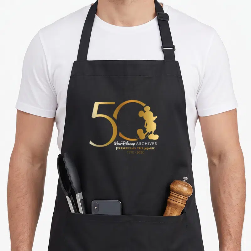 Walt magic apron - Gold 50 with Mickey silhouette and 1970-2020 text — shown on a black cotton canvas apron against a white background