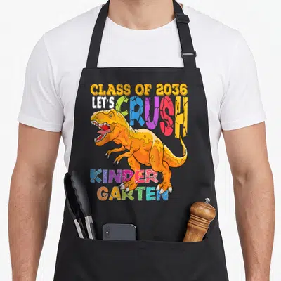 Crush kindergarten school dinosaur apron - Cartoon T-Rex overlapping multicolored text reading CLASS OF 2036 LET'S — shown on a black cotton canvas apron against a white background