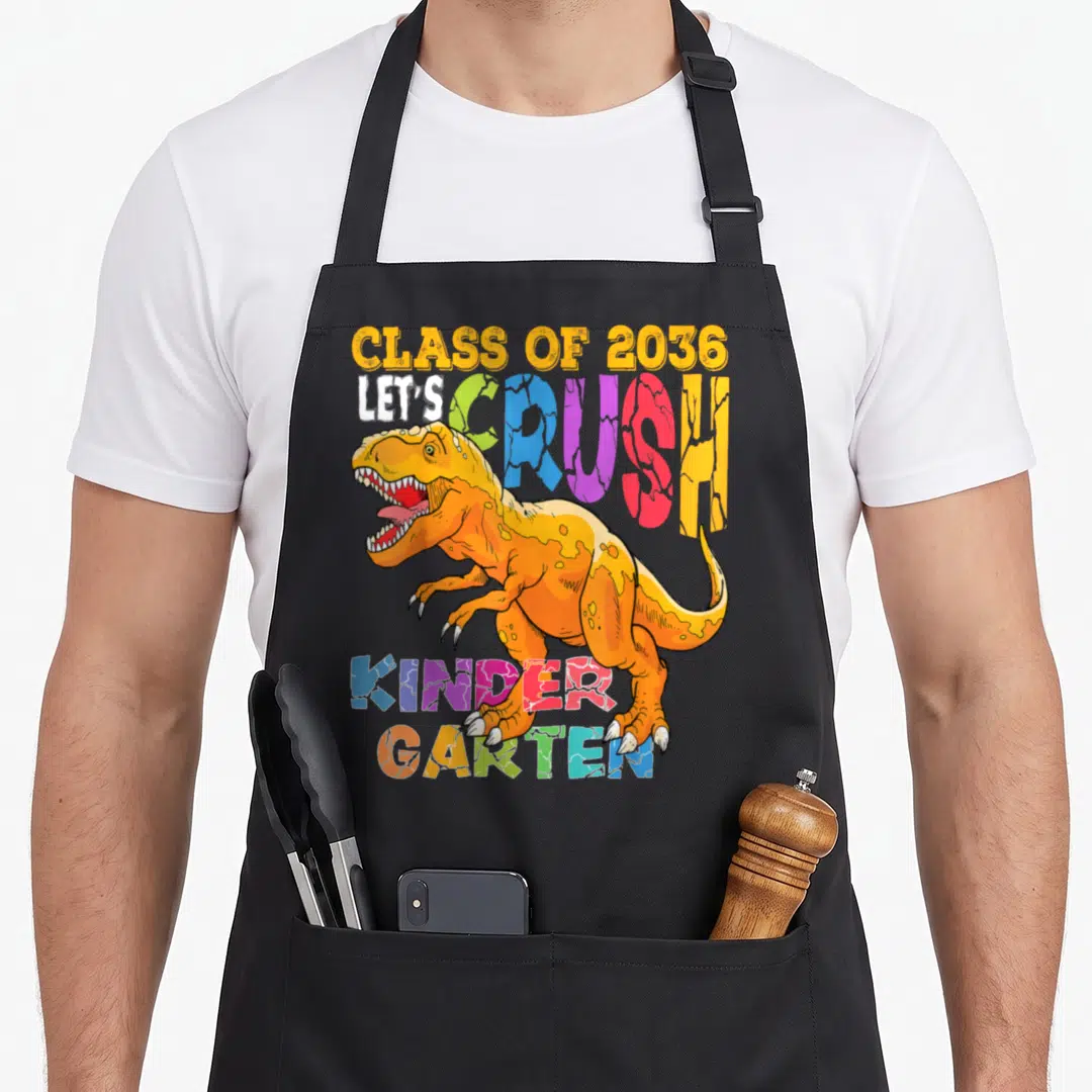 Crush kindergarten school dinosaur apron - Cartoon T-Rex overlapping multicolored text reading CLASS OF 2036 LET'S — shown on a black cotton canvas apron against a white background