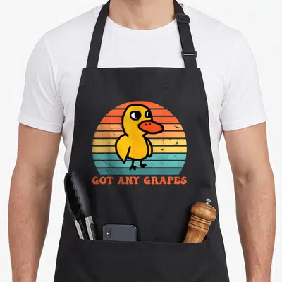 Cute grape duck apron - Yellow cartoon duck on striped orange and teal background with orange text reading GOT ANY — shown on a black cotton canvas apron against a white background