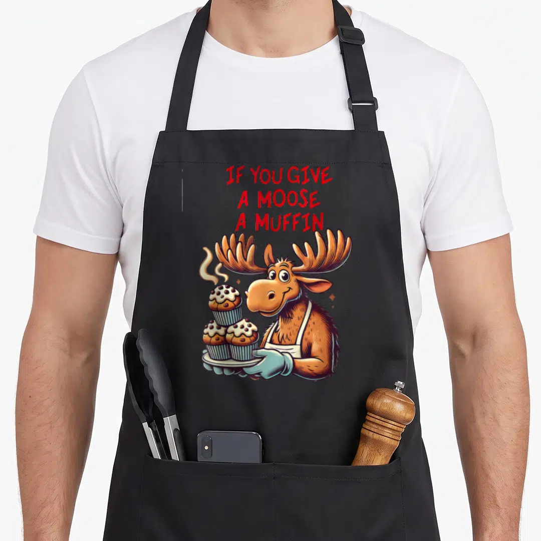 Give muffin man apron - Cartoon moose holding three muffins with red text IF YOU GIVE A MOOSE A MUFFIN — shown on a black cotton canvas apron against a white background
