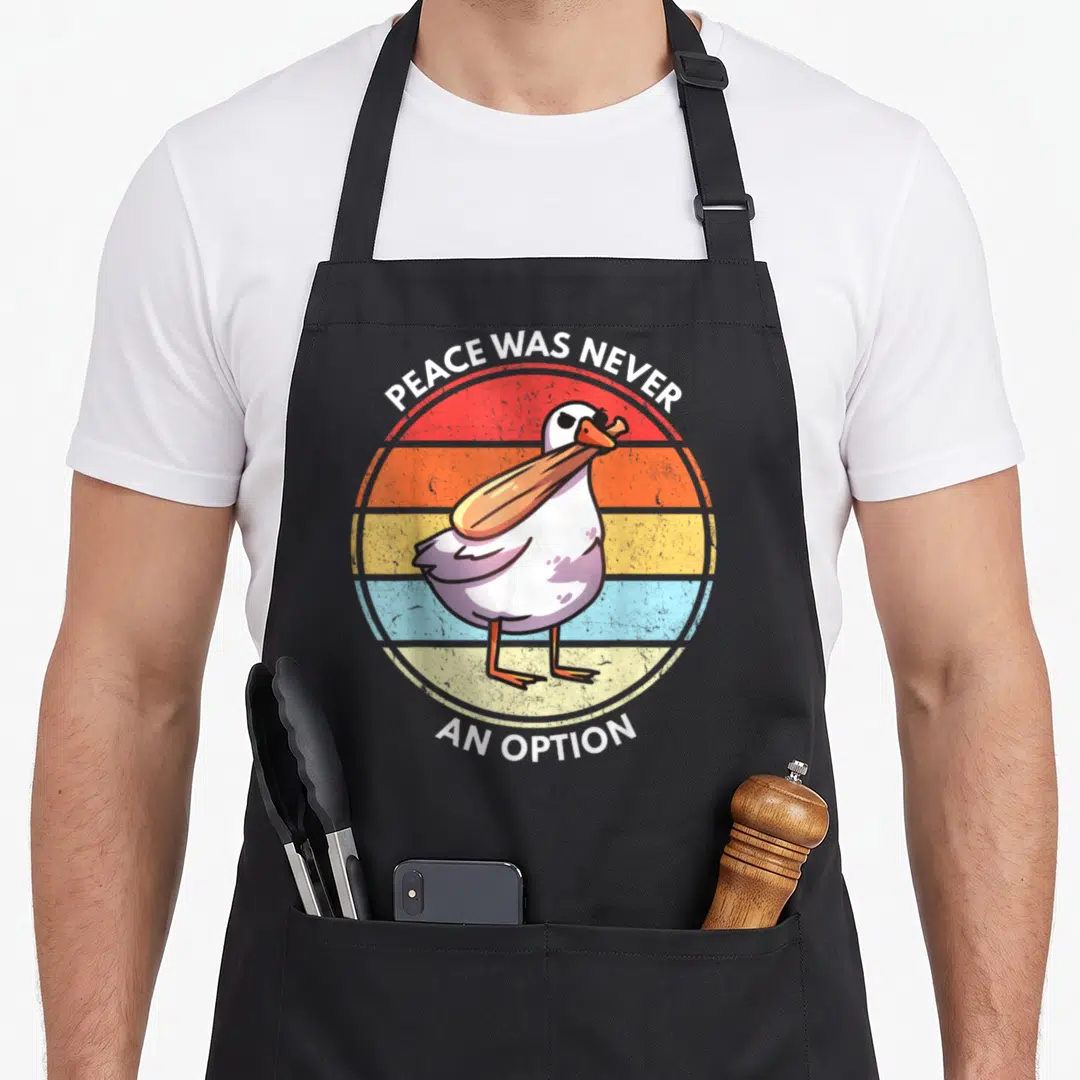 Goose meme apron - Cartoon seagull holding a bat over red orange yellow and blue striped circle with text — shown on a black cotton canvas apron against a white background