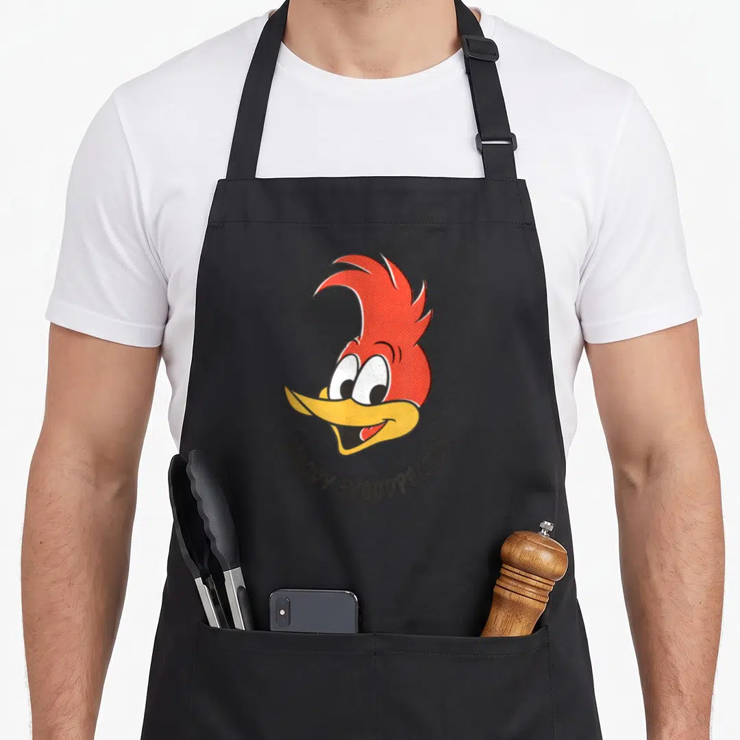 Funny smile woodpecker apron - Woody Woodpecker cartoon head with red crest and curved text in red yellow white grey — shown on a black cotton canvas apron against a white background