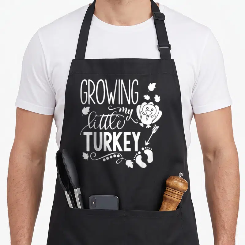 Growing turkey thanksgiving apron - White stacked text GROWING my little TURKEY with cartoon bird and baby footprints — shown on a black cotton canvas apron against a white background