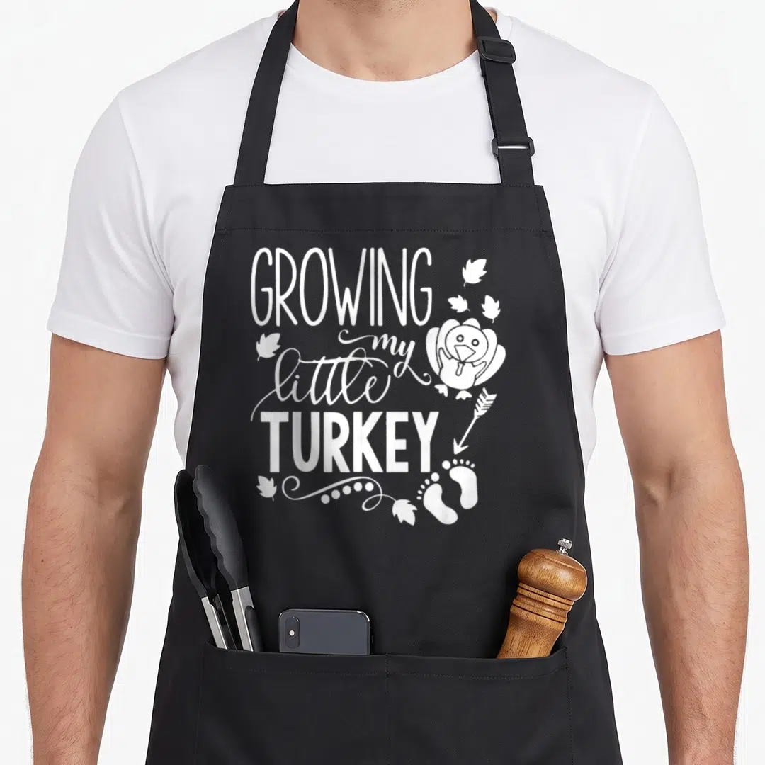 Growing turkey thanksgiving apron - White stacked text GROWING my little TURKEY with cartoon bird and baby footprints — shown on a black cotton canvas apron against a white background