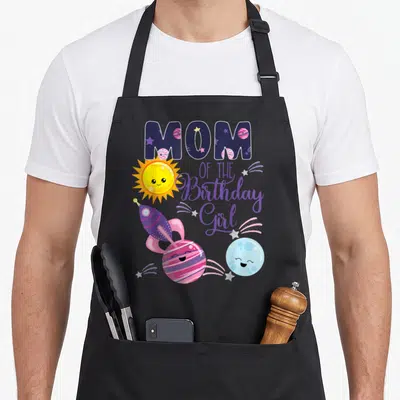 Outer space theme birthday apron - MOM OF THE Birthday Girl text with cartoon sun, rocket, pink planet, and blue moon — shown on a black cotton canvas apron against a white background