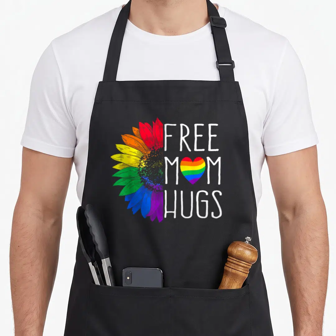 Free sunflower apron - Rainbow sunflower next to stacked FREE MOM HUGS text with rainbow heart replacing O in MOM — shown on a black cotton canvas apron against a white background