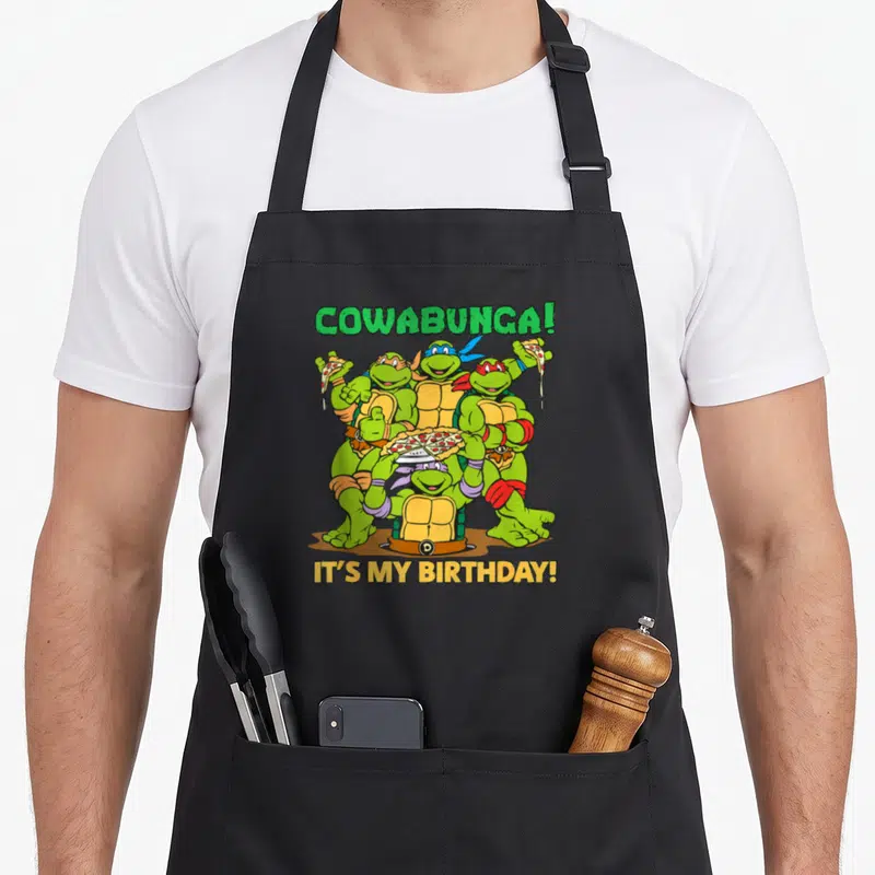 Cowabunga birthday apron - Four turtles holding pizza with COWABUNGA! IT'S MY BIRTHDAY! text — shown on a black cotton canvas apron against a white background