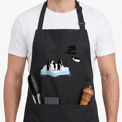 Funny fan penguin aprons - Cartoon penguin jumping in air near three standing birds on blue ice with bold text — shown on a black cotton canvas apron against a white background