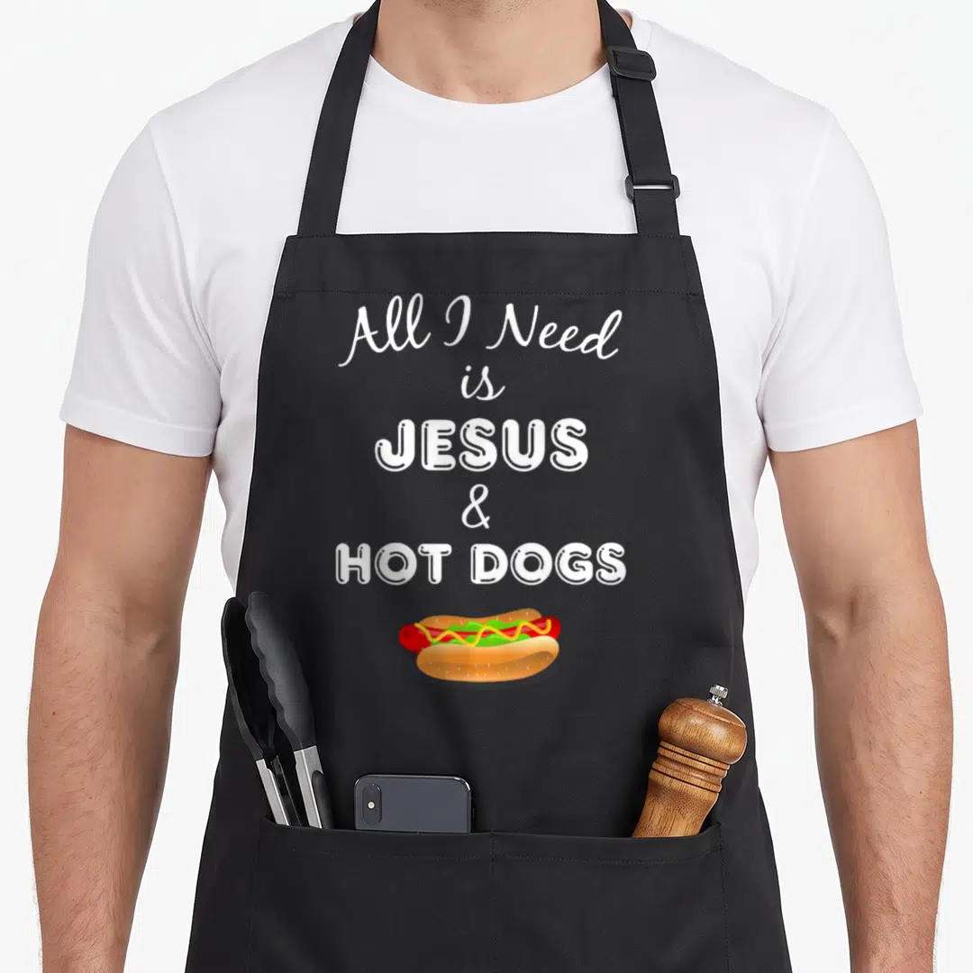 Jesus hot dog apron - Stacked white text reading All I Need is JESUS & HOT DOGS above a cartoon hot dog with mustard — shown on a black cotton canvas apron against a white background
