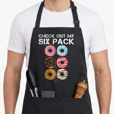 Six donut apron - CHECK OUT MY SIX PACK text above six colorful glazed donuts with sprinkles and star toppings in two — shown on a black cotton canvas apron against a white background