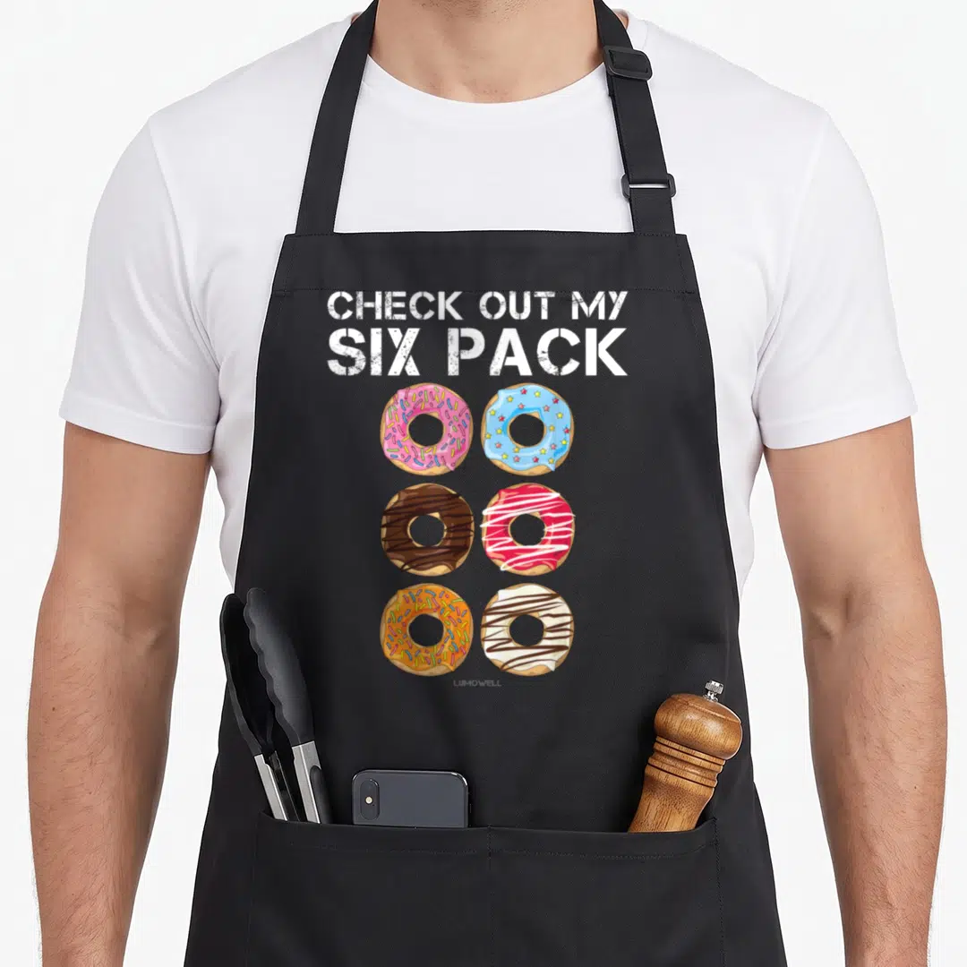 Six donut apron - CHECK OUT MY SIX PACK text above six colorful glazed donuts with sprinkles and star toppings in two — shown on a black cotton canvas apron against a white background