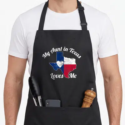 Aunt texans apron - Texas map shape with blue white and red sections and a central heart surrounded by white script text — shown on a black cotton canvas apron against a white background