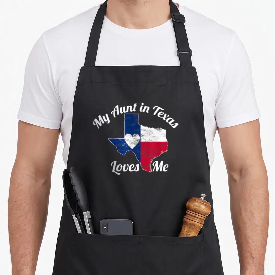 Aunt texans apron - Texas map shape with blue white and red sections and a central heart surrounded by white script text — shown on a black cotton canvas apron against a white background