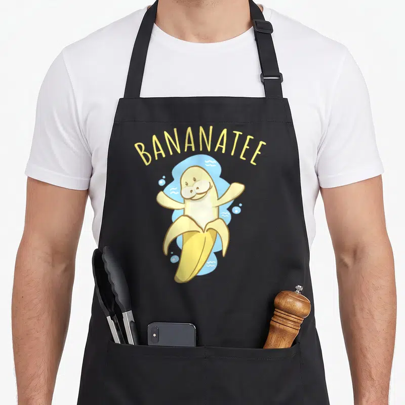 Bananatee fruit apron - Yellow manatee banana hybrid character under arched BANANATEE text on blue splash — shown on a black cotton canvas apron against a white background