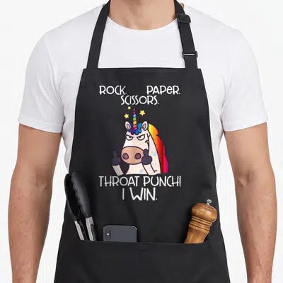 Win paper apron - Cartoon unicorn giving thumbs up with Rock Paper Scissors Throat Punch text and stars — shown on a black cotton canvas apron against a white background