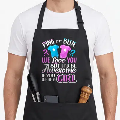 Funny blue apron or apron - Pink and blue onesies with stars flanked by question marks and stacked gender reveal text — shown on a black cotton canvas apron against a white background