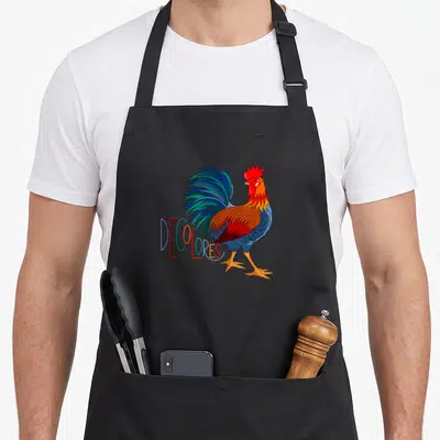 Gsallicat rooster apron - Colorful cartoon rooster with DECOLORES text in red blue green orange yellow — shown on a black cotton canvas apron against a white background