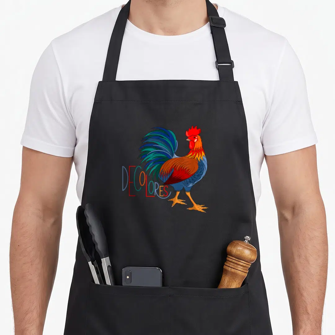 Gsallicat rooster apron - Colorful cartoon rooster with DECOLORES text in red blue green orange yellow — shown on a black cotton canvas apron against a white background