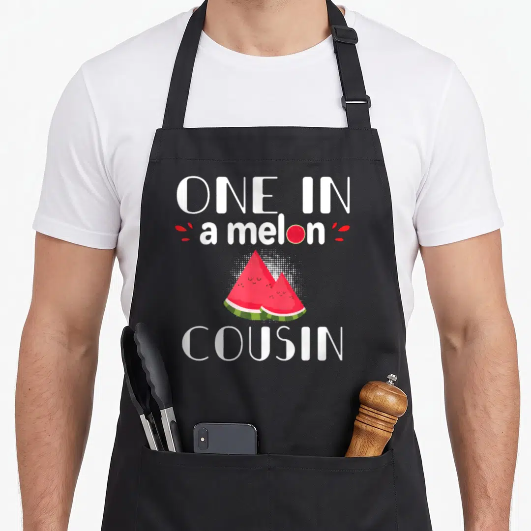 Cousin watermelon apron - One In a Melon Cousin text with two red cartoon watermelon slices — shown on a black cotton canvas apron against a white background