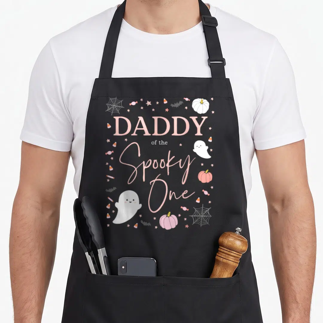 Daddy funny halloween apron - Pink and white text Daddy of the Spooky One with ghost, pumpkins, candy corn and stars — shown on a black cotton canvas apron against a white background